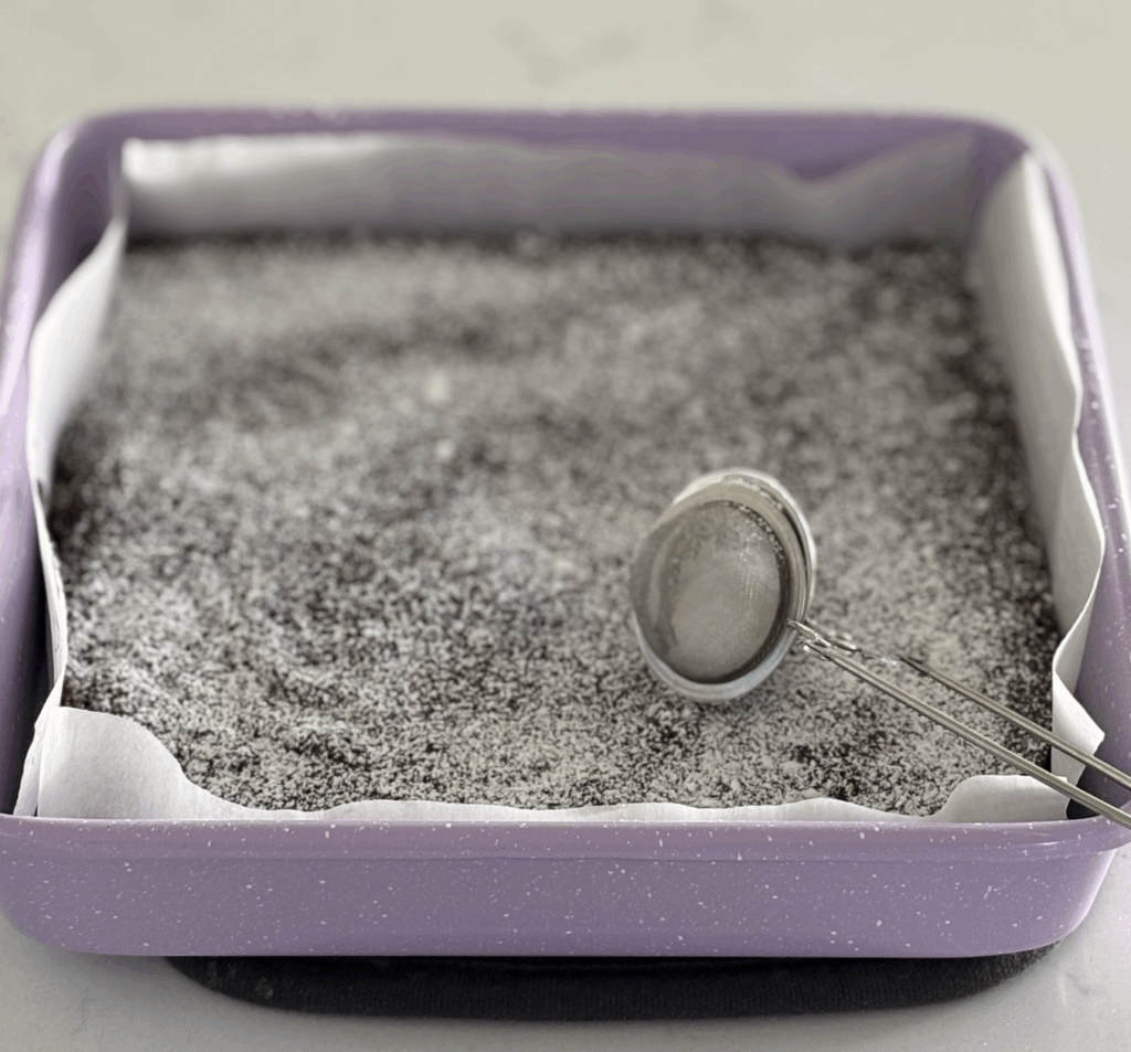 Freshly baked egg-free, dairy-free brownies in a parchment-lined pan dusted with powdered sugar, with a stainless steel tea infuser used for sprinkling.