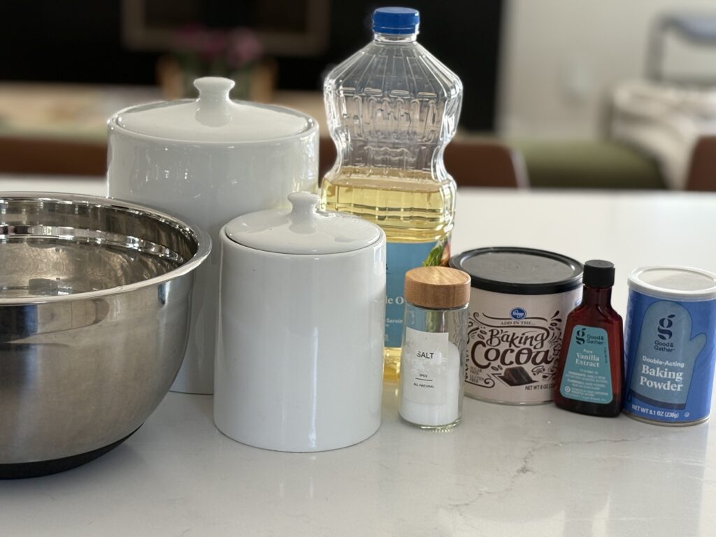 Ingredients for egg-free, dairy-free brownies displayed on a kitchen counter, including vegetable oil, cocoa powder, vanilla extract, baking powder, salt, sugar, and mixing bowls.