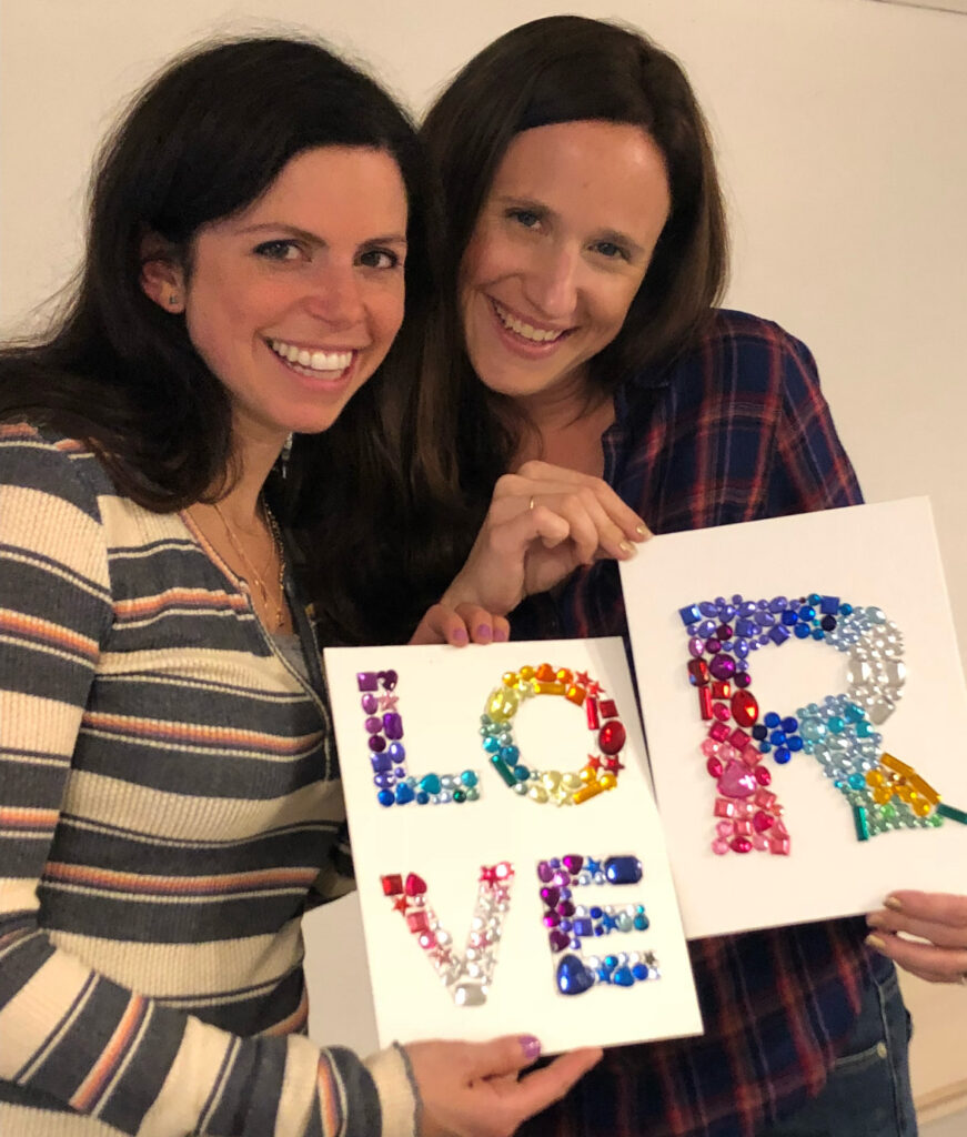 Calie and Dara, founders of The Tortured Sugar Department, smiling and holding colorful artwork that spells out the word LOVE.