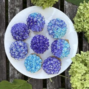 Purple and blue hydrangea cupcakes arranged on a white plate surrounded by green hydrangea leaves, made egg-free and nut-free.