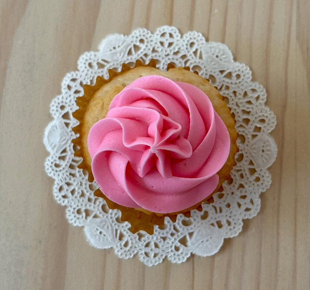 Egg-free and nut-free vanilla cupcake topped with pink buttercream frosting on a lace doily.