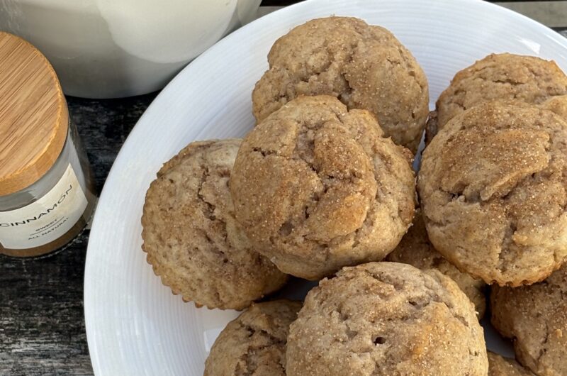 Overhead view of golden brown cinnamon muffins topped with cinnamon sugar on a white plate, next to a cinnamon spice jar.